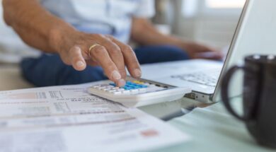 Serious elder senior grey-haired grandpa using calculator counting bank loan payment holding papers at home, focused mature old man doing paperwork calculating bills managing finances sit on sofa